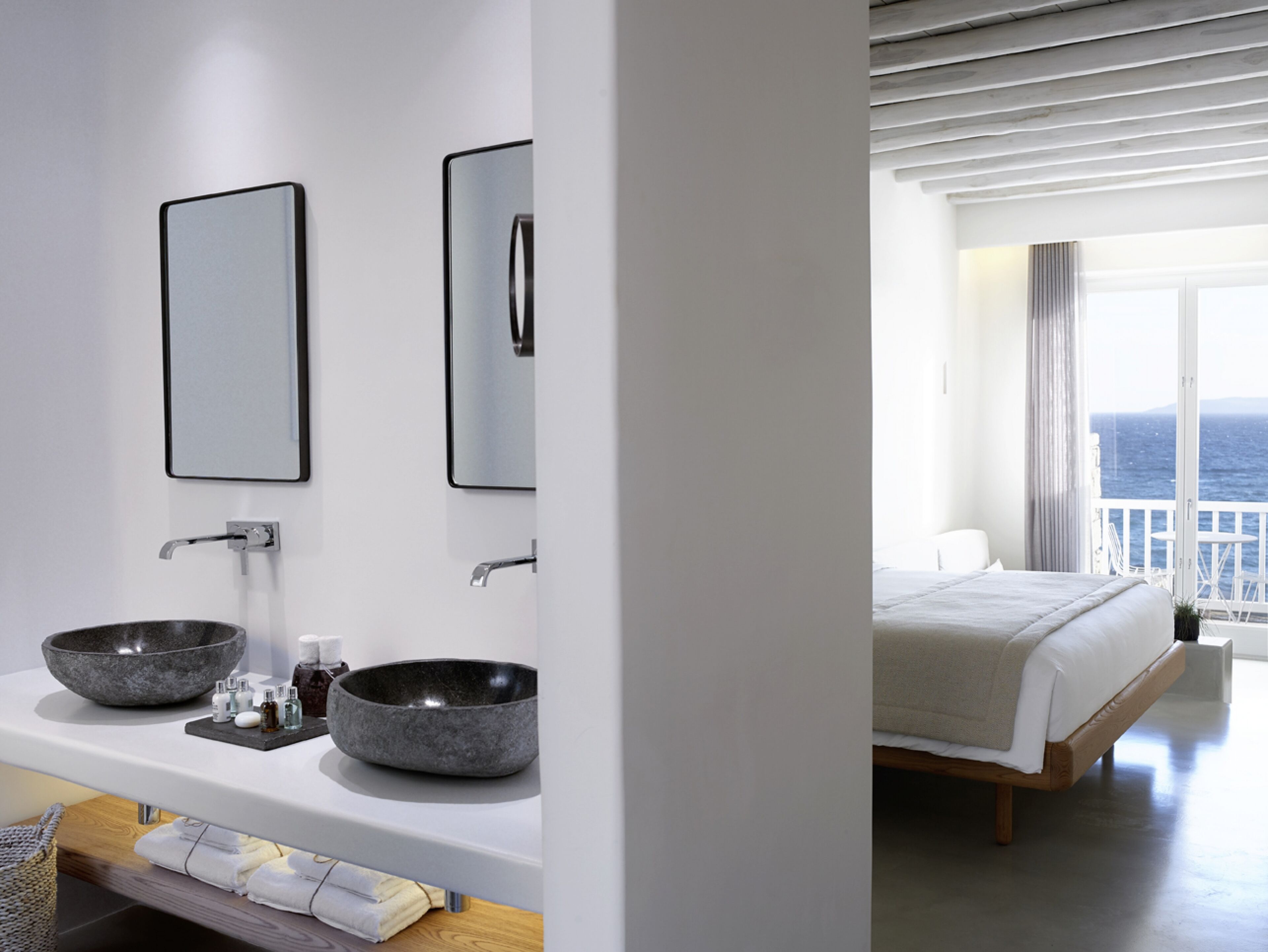 A modern bathroom with two large stone sinks, a mirror, and a wooden vanity. The room has a large window with a view of the sea.