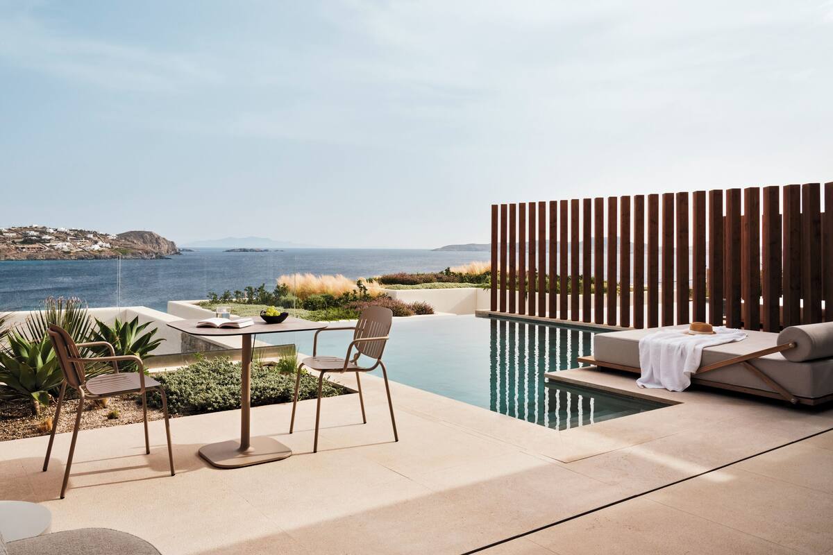 Zona de piscina exterior con vista al mar, una mesa con sillas y una valla de madera.