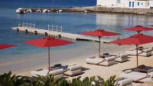 A beach with sun loungers and red umbrellas, a pier, and a white building with blue doors. - The Coast Bill&Coo - Adults Only (Mykonos)