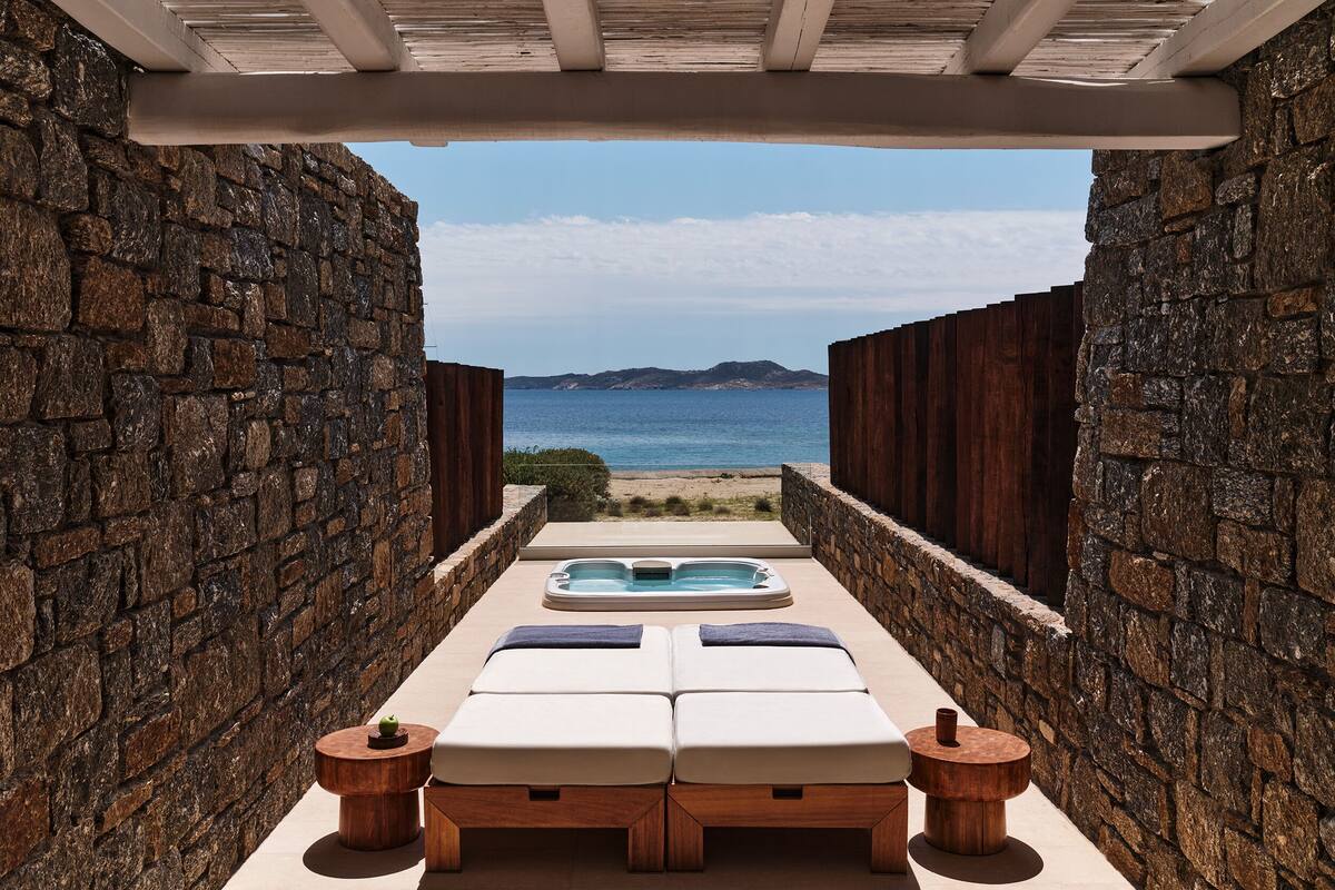 Un espacio al aire libre con paredes de piedra, con hidromasaje y sillones reclinables, con vista a una playa y al mar.