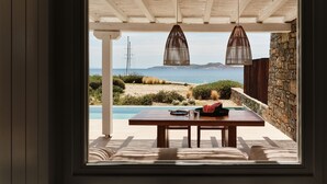 A view from a window overlooking a pool area with a wooden deck, two hanging lamps, and a stone wall. - The Coast Bill&Coo - Adults Only (Mykonos)
