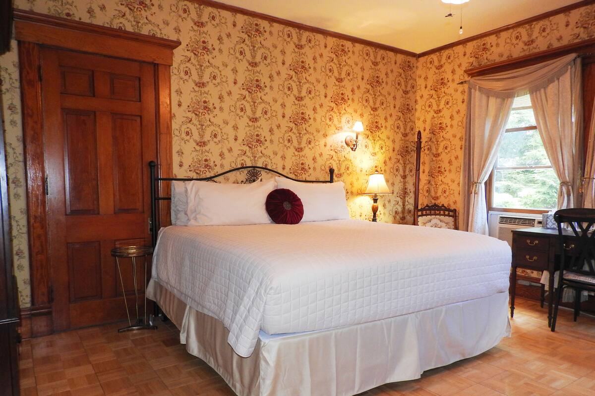 A bedroom with a large bed, wooden door, floral wallpaper, and a desk with a chair.