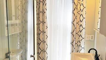 Un baño con cabina de ducha de vidrio, inodoro, lavamanos y ventana con cortinas estampadas.