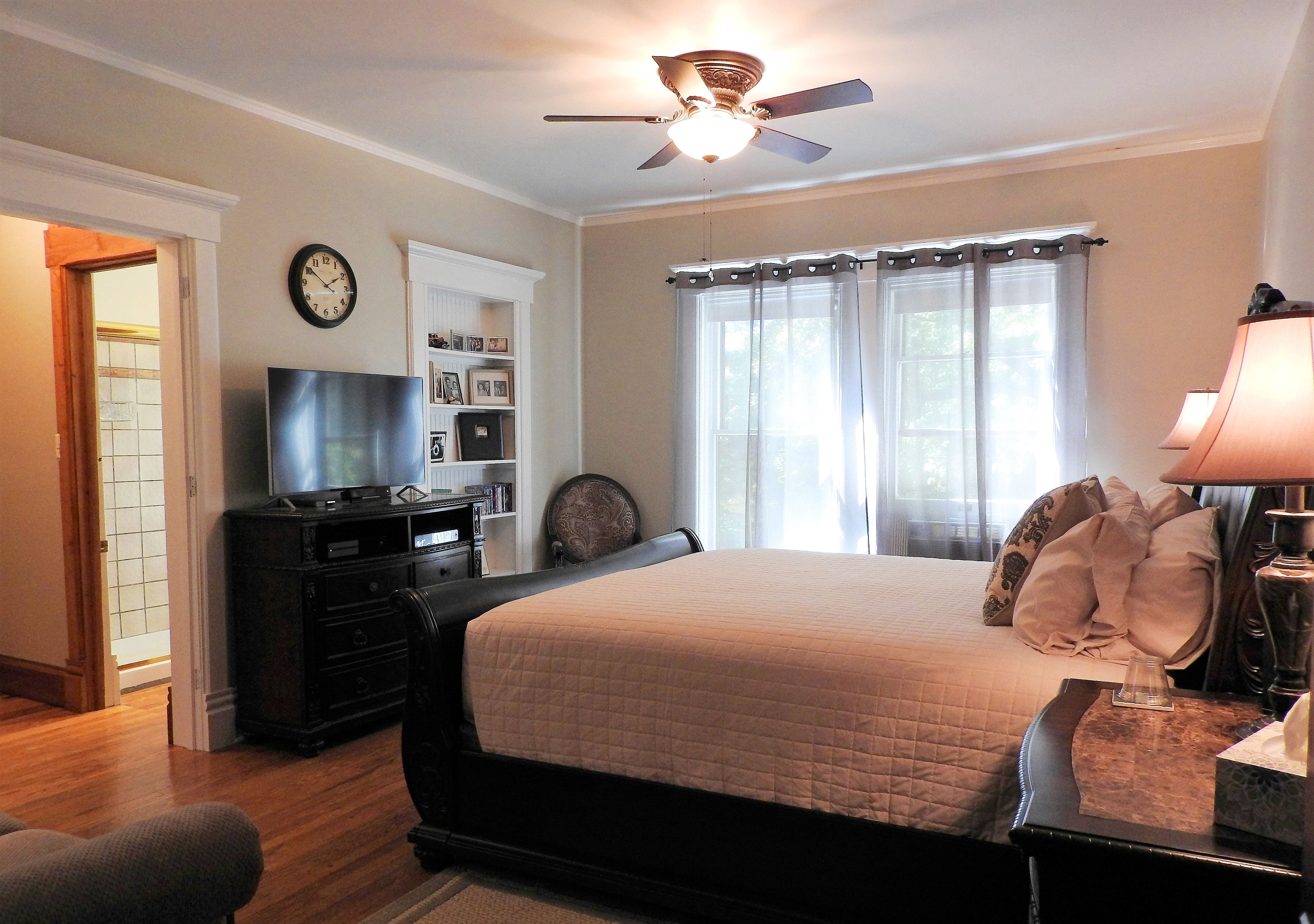 A bedroom with a bed, a ceiling fan, a TV, a bookshelf, and a window with curtains.