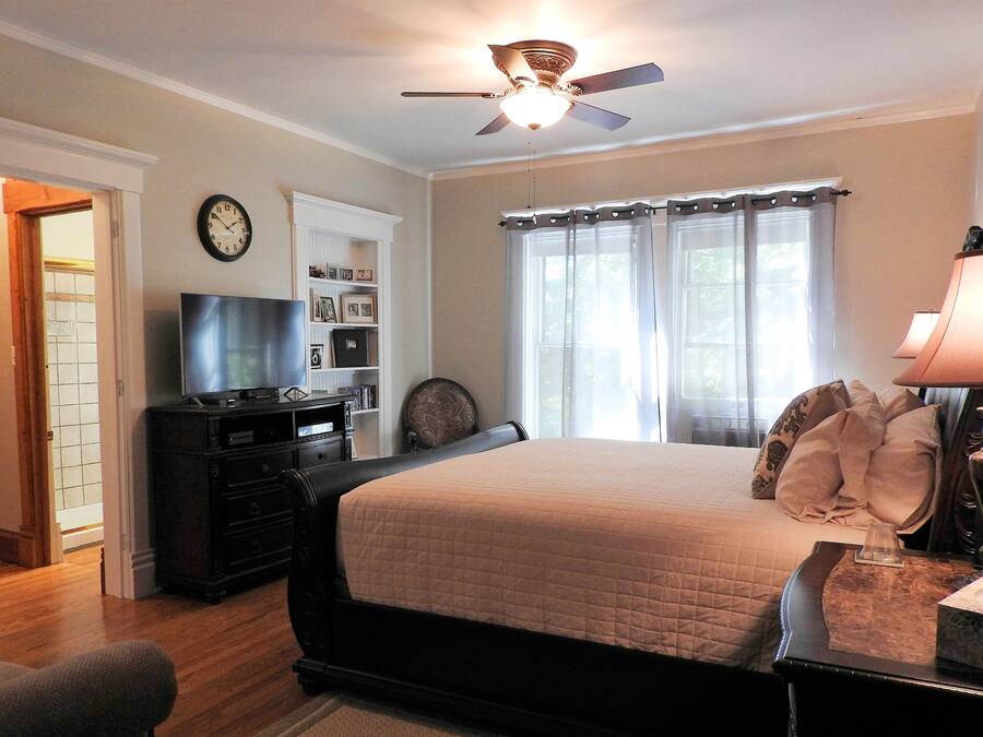 A bedroom with a bed, a ceiling fan, a TV, a bookshelf, and a window with curtains.