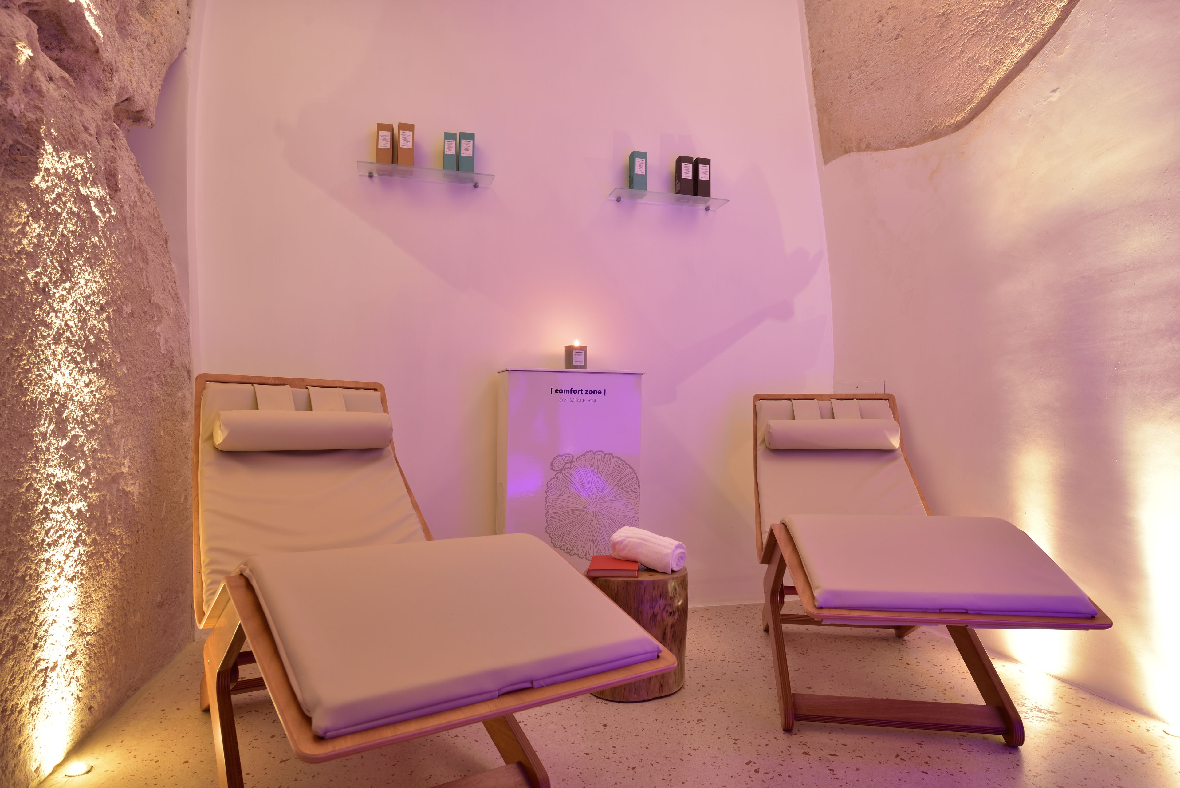 A spa room with two wooden loungers, a small wooden stool with a towel, and a wall-mounted shelf with items.