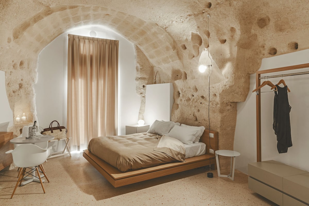 A bedroom with a stone wall, a bed, a bedside table, a chair, and a hanging coat rack.
