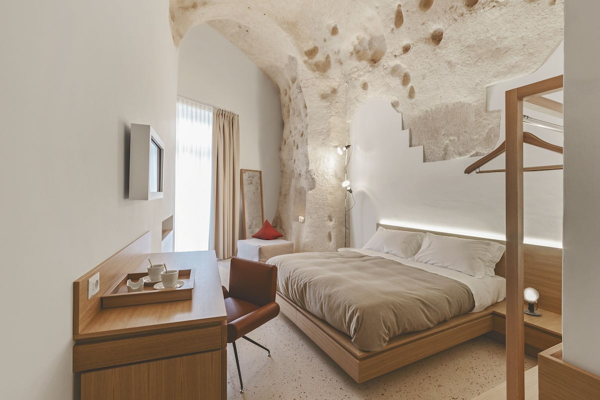 A modern bedroom with a wooden desk, a bed with a beige bedspread, a red pillow, and a stone wall.
