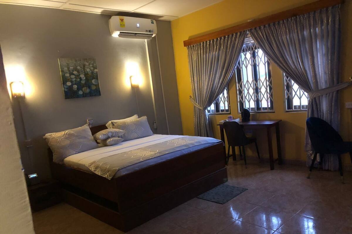 A bedroom with a bed, a desk, and a chair. There is a window with curtains and a painting on the wall.