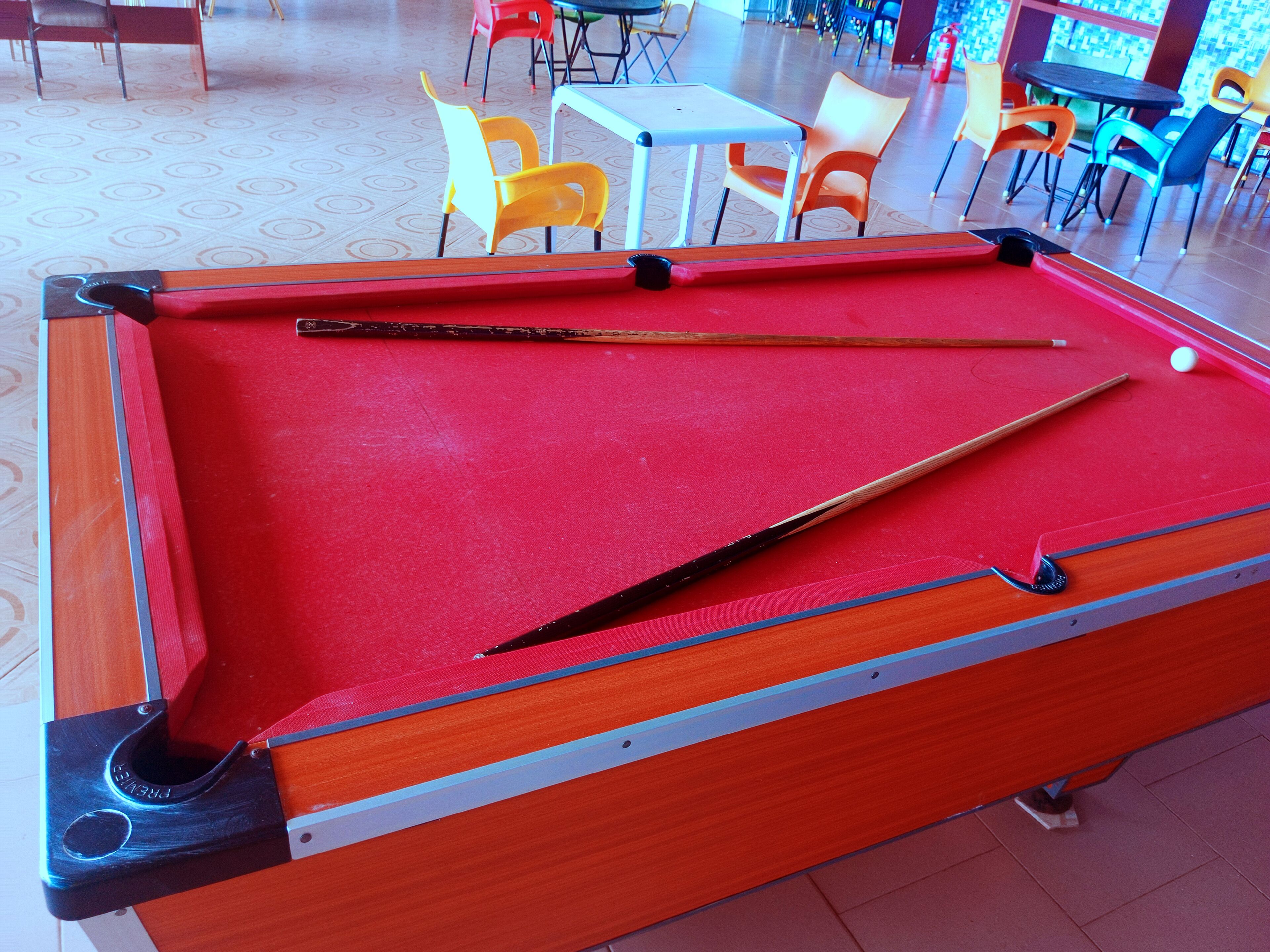 Una mesa de billar con superficie de fieltro rojo, rodeada de sillas y mesas en una sala con suelo estampado.