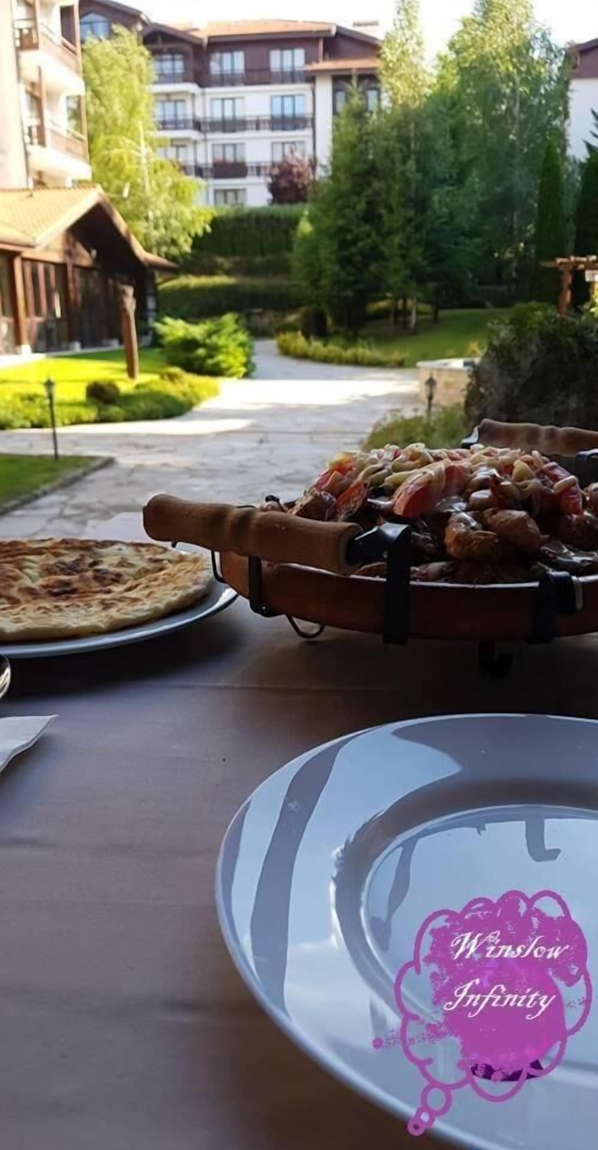 Una mesa de comedor con pizza y brochetas de carne, frente a un paisaje urbano con edificios y vegetación.