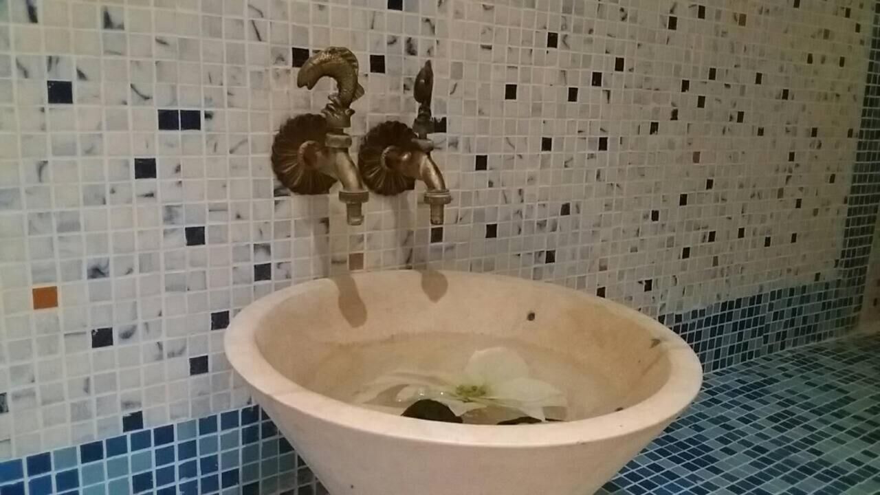 A bathroom with a large, round, beige sink and two ornate faucets. The walls are tiled with a mosaic pattern.