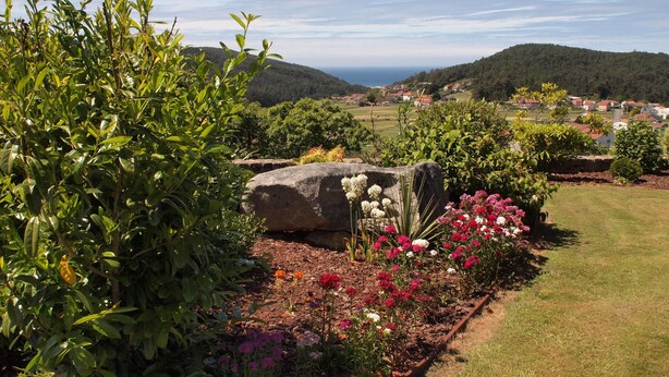 A garden with various plants and flowers, a large rock, and a view of a town in the background. - Hotel Rústico A Torre de Laxe (Laxe)