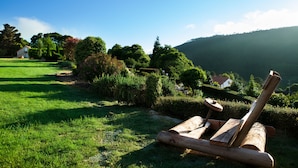 A wooden log chair in a lush garden with a clear view of a house and hills in the background. - Hotel Rústico A Torre de Laxe (Laxe)