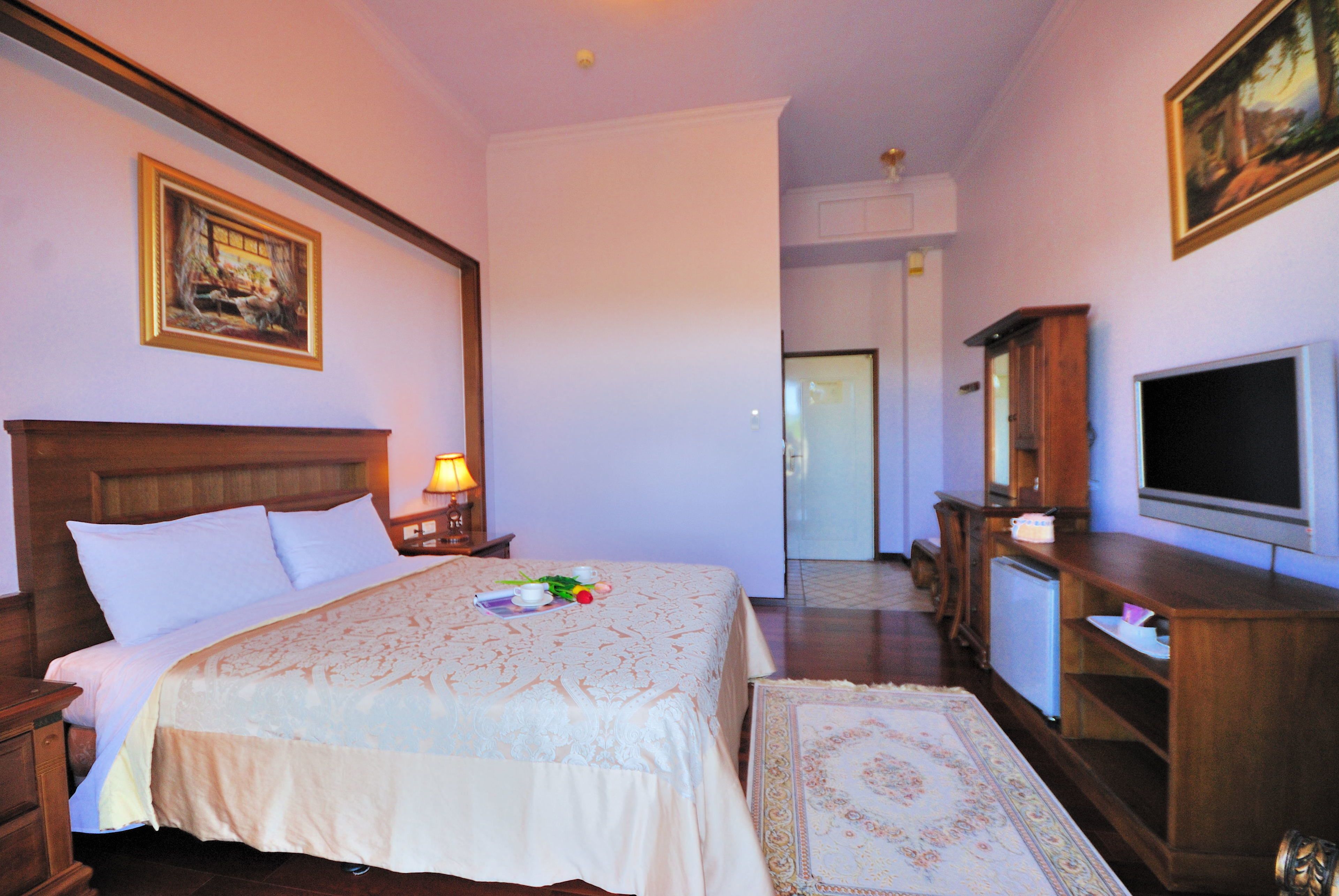 A hotel room with a large bed, a TV, a wooden cabinet, and a desk with a lamp.