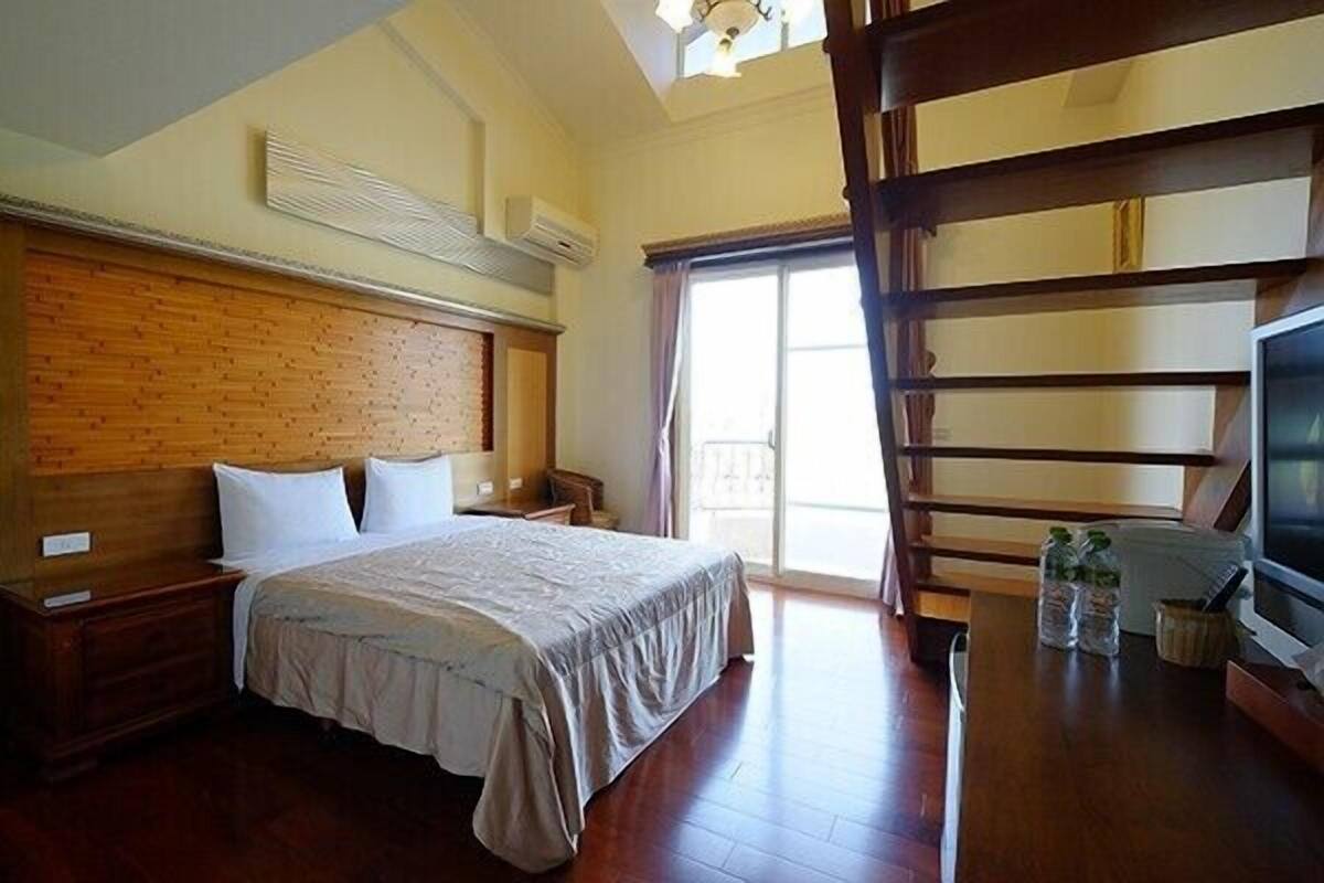 Habitación de hotel con cabecera de madera, cama con colcha gris, escritorio de madera con televisor y ventana con cortinas.