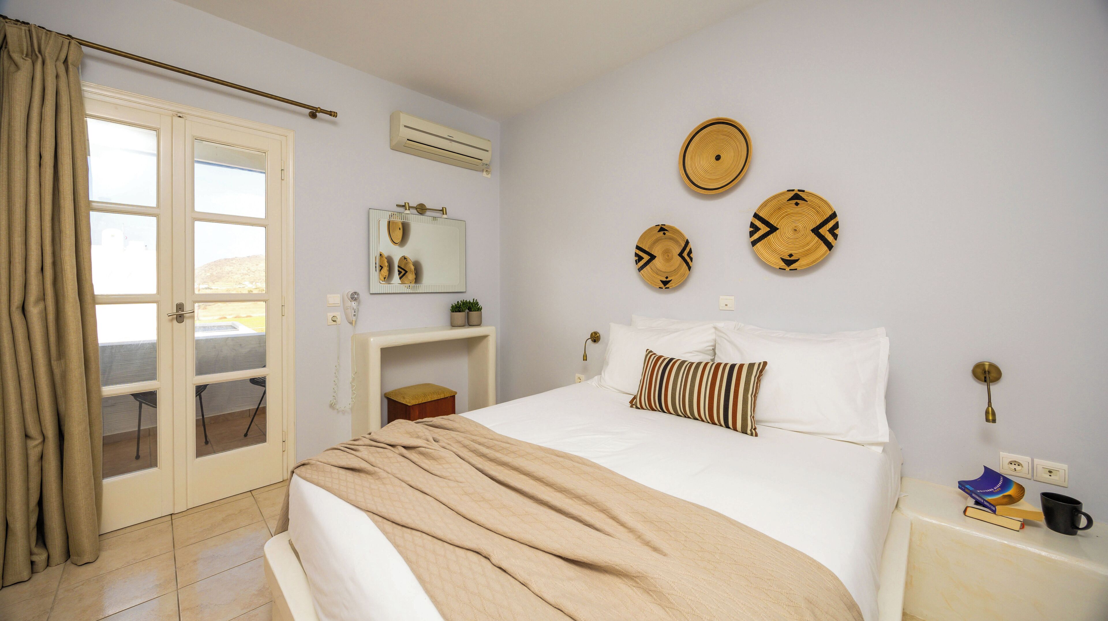 A bedroom with a bed, a bedside table with a lamp, a wall-mounted mirror, and a window with curtains.
