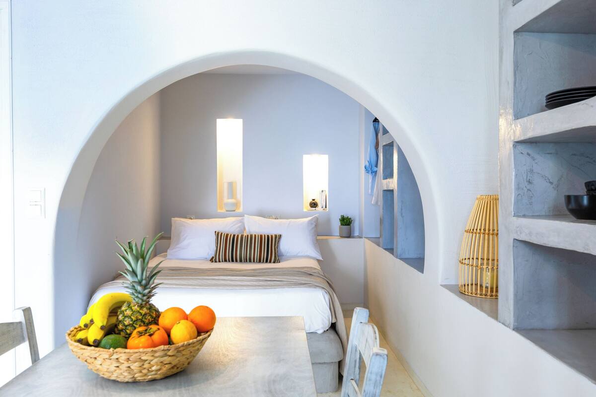 A modern bedroom with an arched doorway, a bed with white linens, a bedside table with a fruit bowl, and a small potted plant.