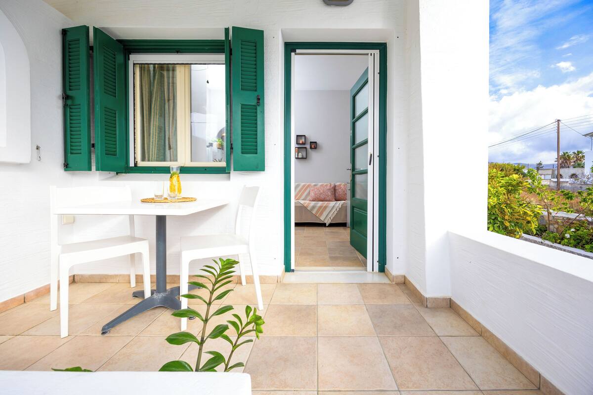 A balcony with a white table and chairs, a potted plant, and a view of a garden and buildings.