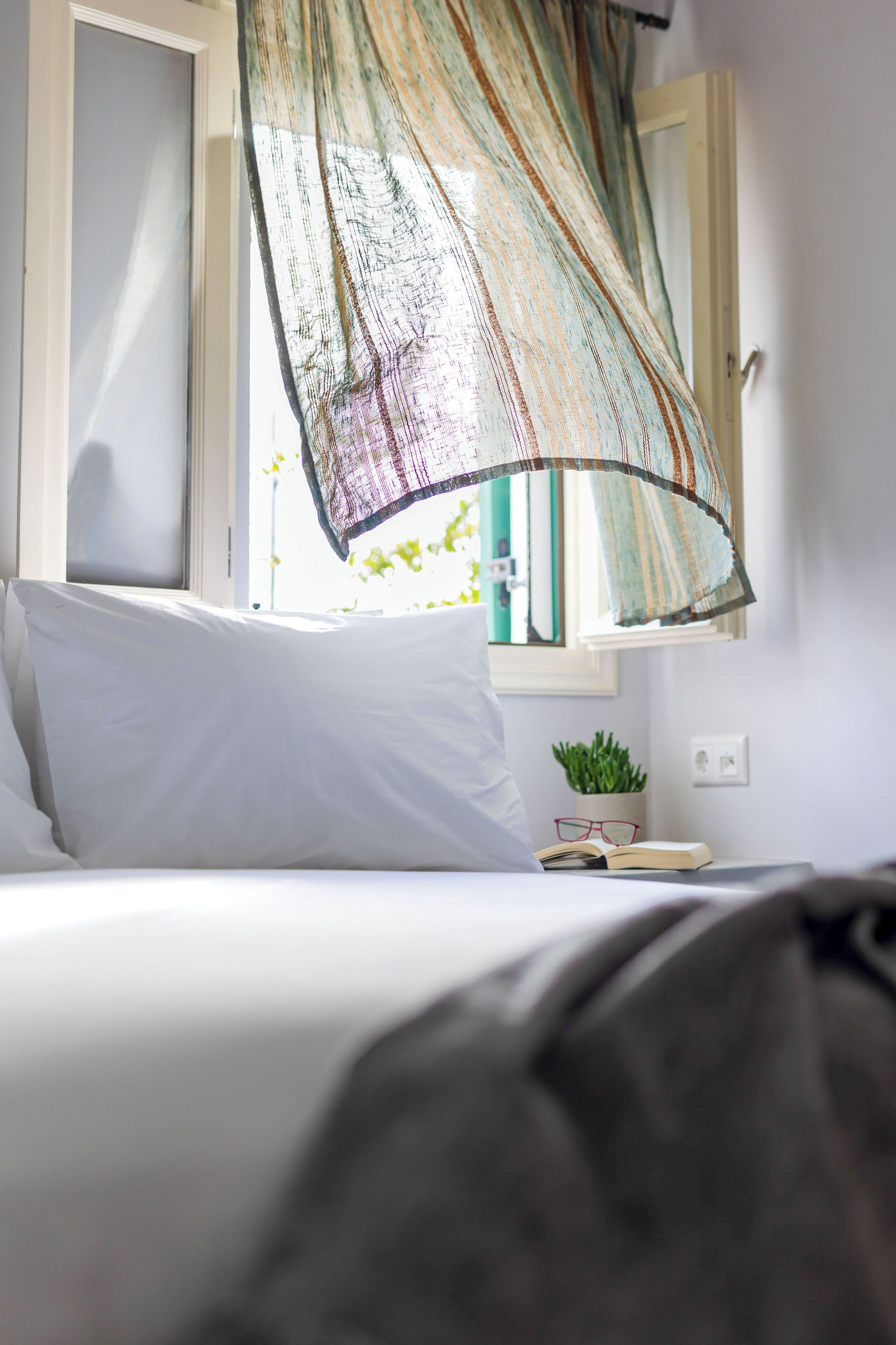 A bed with white bedding, a window with a view of a plant and books, and a hanging curtain.