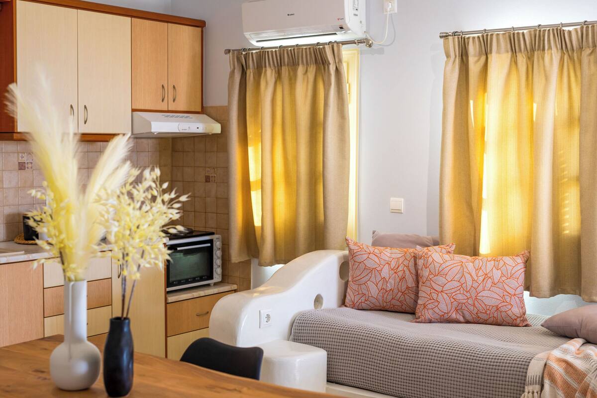 A compact living space with a kitchenette, a bed with patterned pillows, and a dining area with a vase of flowers.