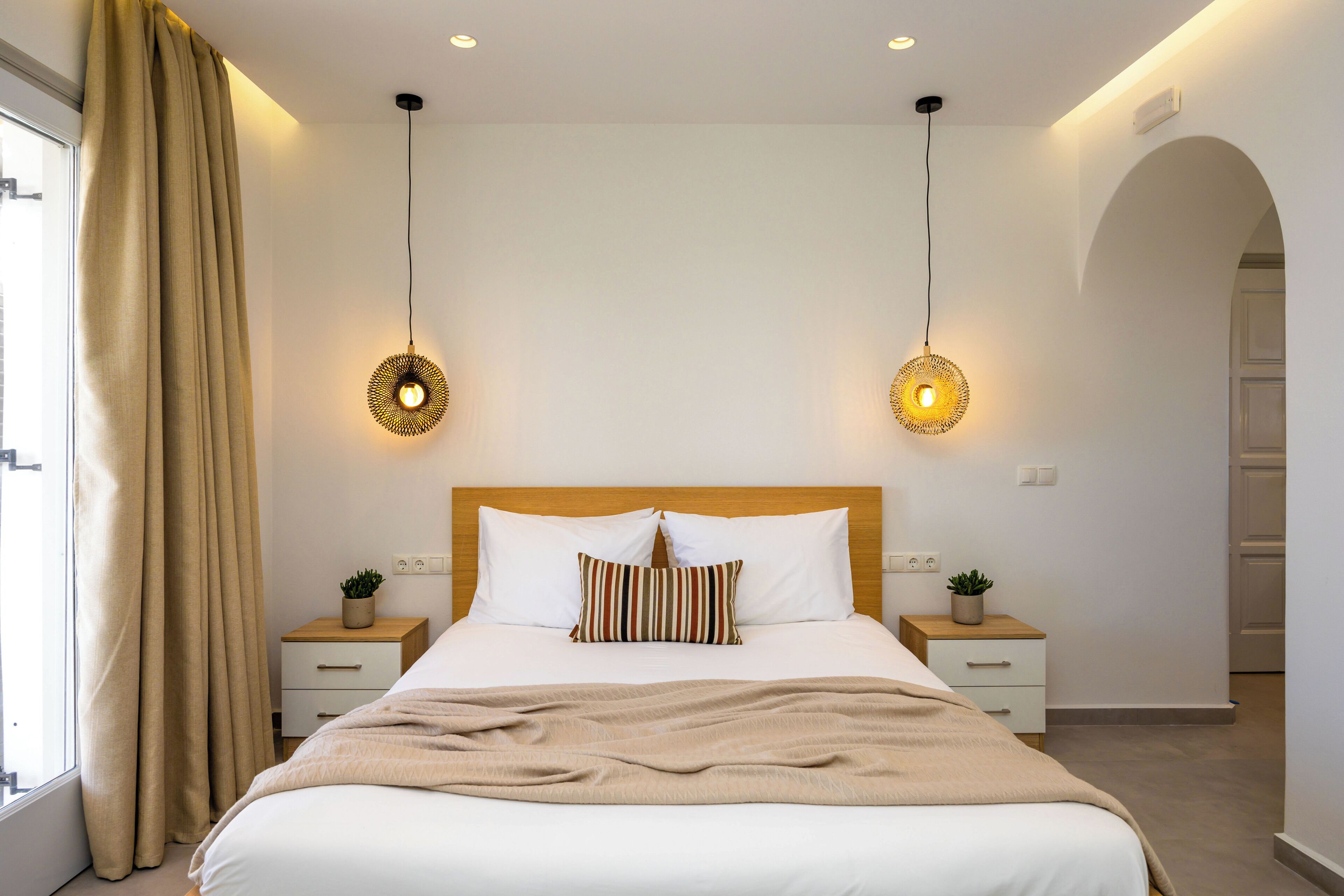A modern bedroom with a large bed, two bedside tables with plants, a wooden headboard, and a large window with curtains.