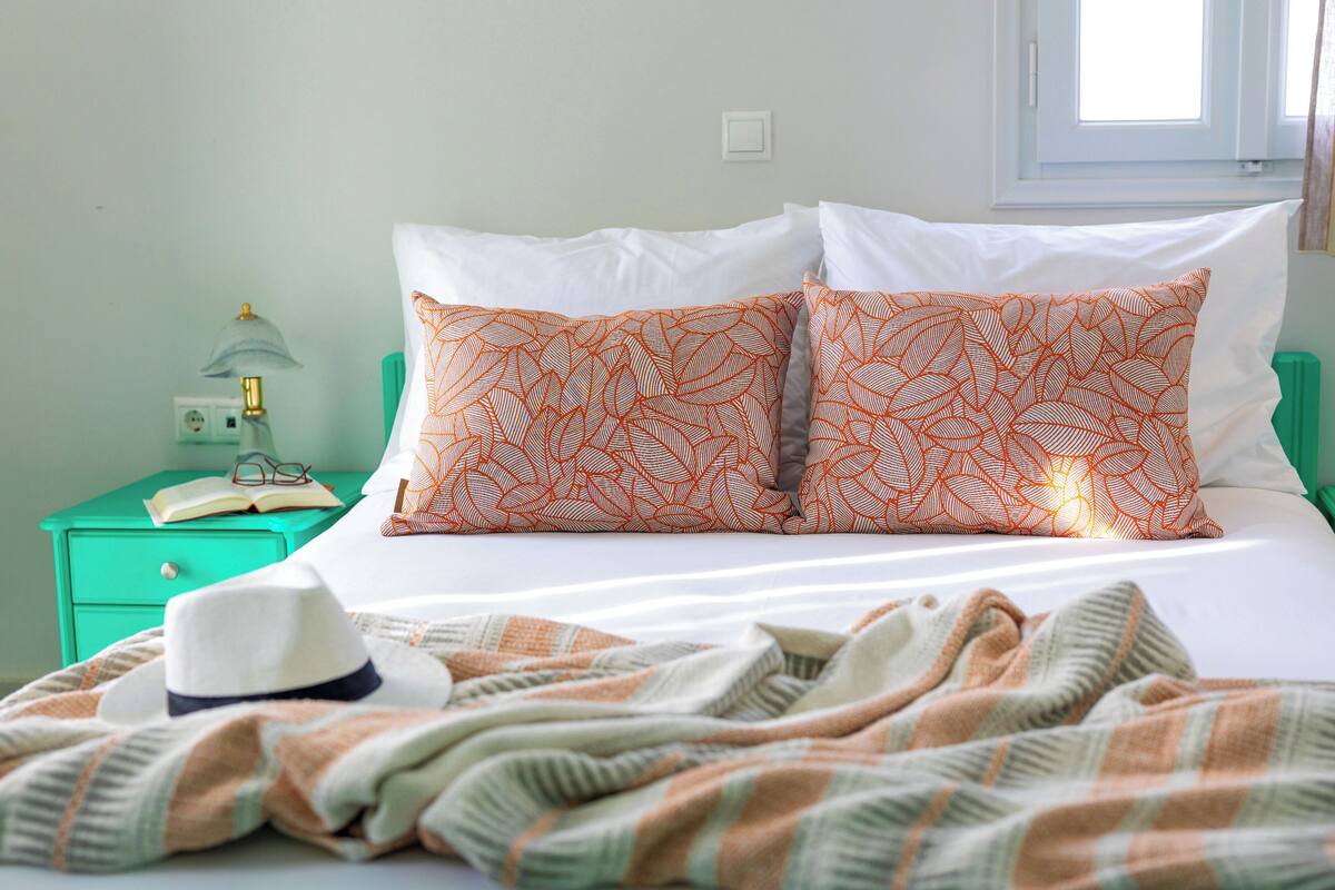 A neatly made bed with white and orange patterned pillows, a green nightstand with a lamp, and a plaid blanket.