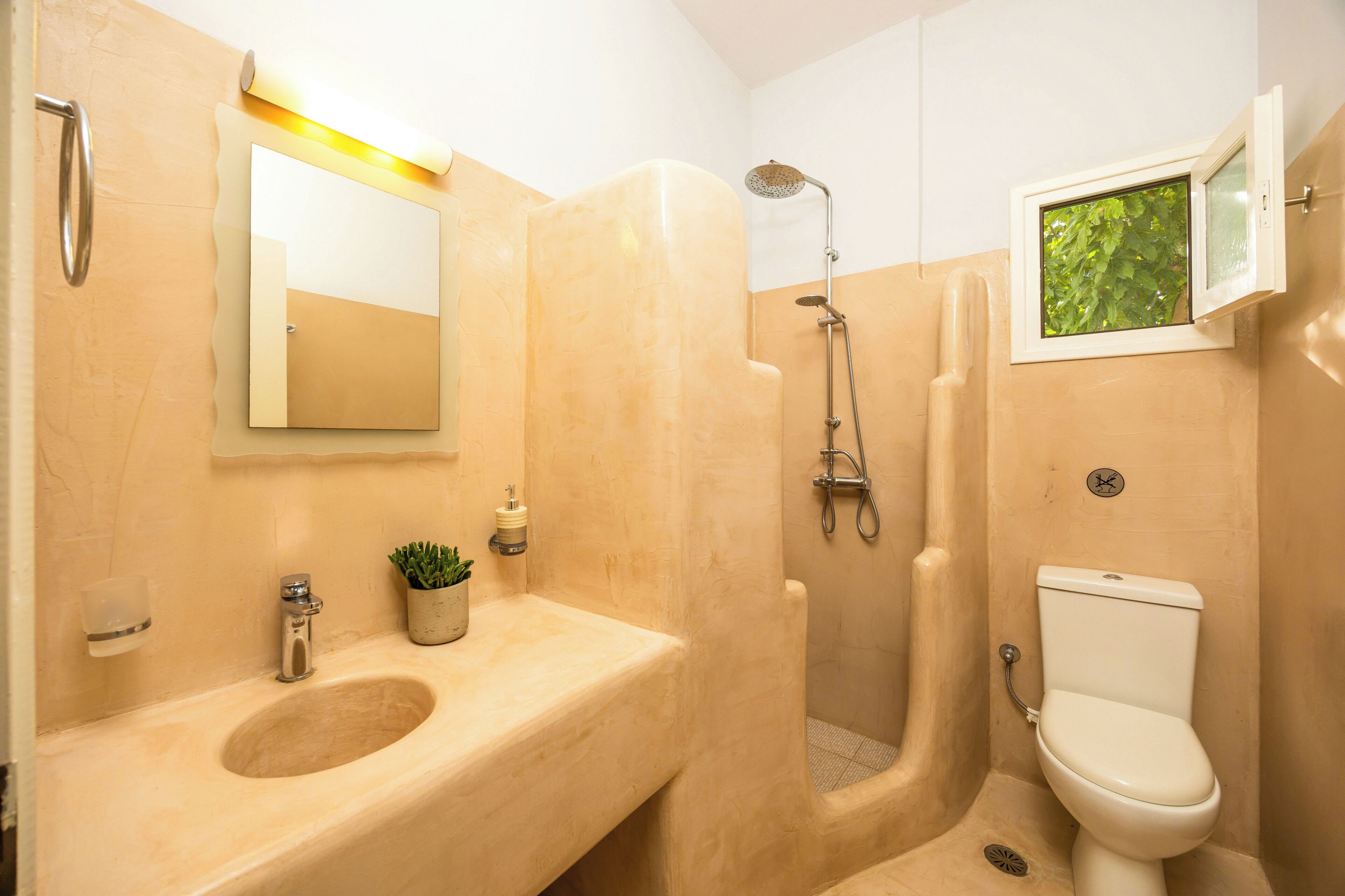 Une salle de bain équipée d’un lavabo, d’un miroir, de toilettes, d’une douche et d’une fenêtre donnant sur un espace verdoyant.