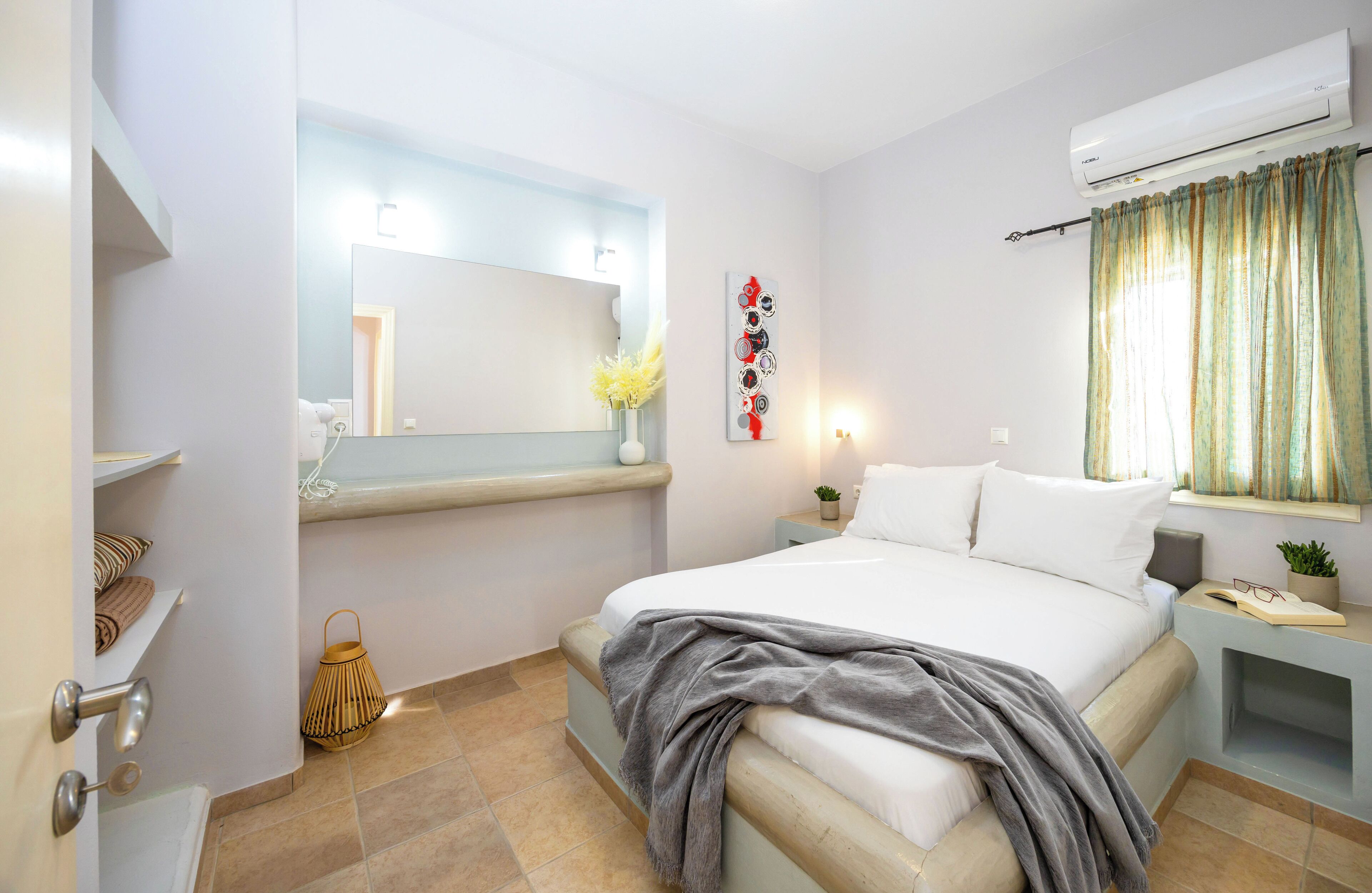 A modern bedroom with a large bed, a bedside table with a lamp, a wall-mounted air conditioner, and a window with curtains.