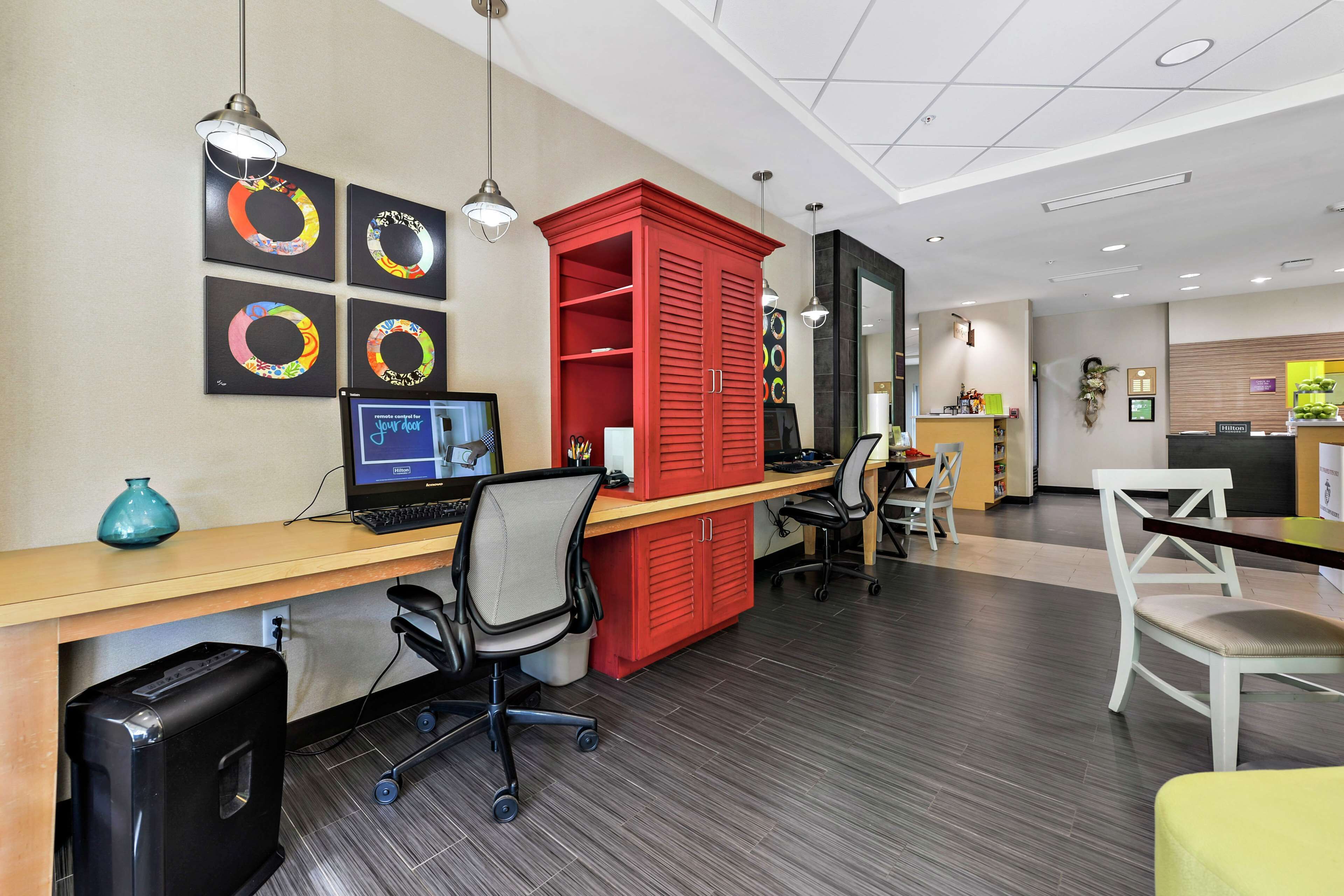 A modern office space with a desk, computer, and office chairs. There are framed artworks on the wall and a red cabinet.