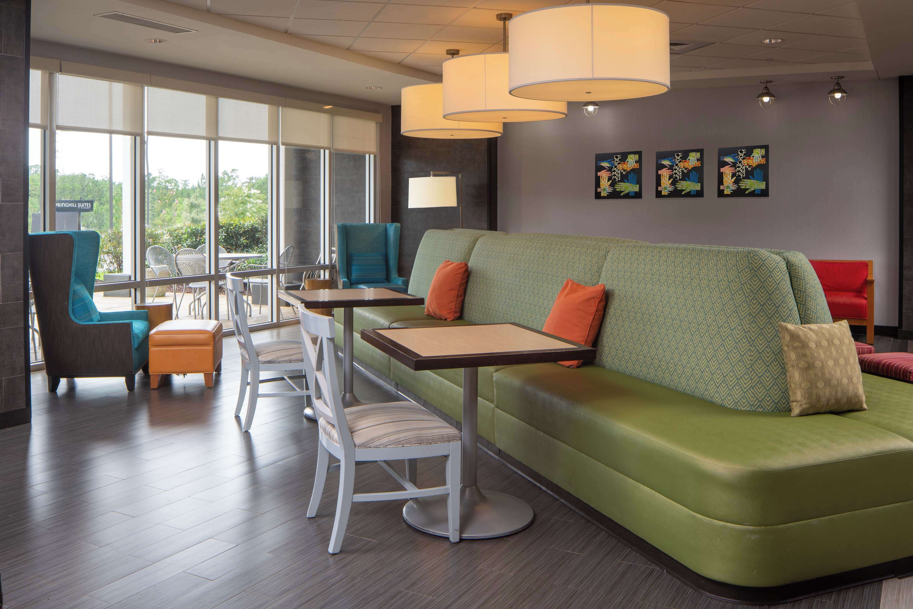 A modern lounge area with a green sofa, white chairs, and tables. There are large windows providing a view of greenery outside.