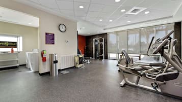 A gym with cardio machines, a wall clock, and a window with a view of palm trees.