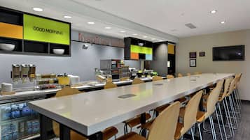 A modern breakfast area with a long communal table, seating, and a refrigerator stocked with beverages.