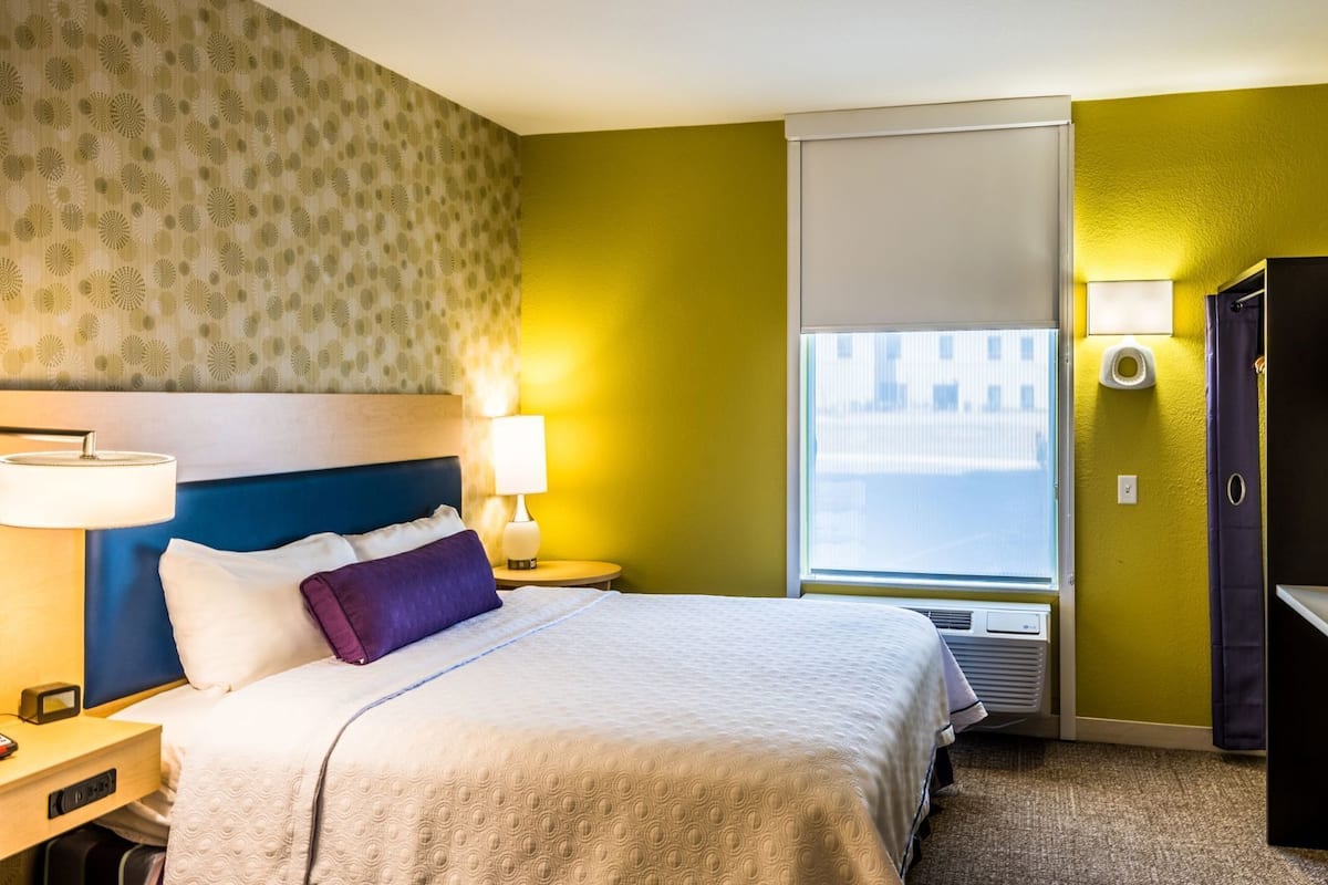 Hotellihuone, jossa on sänky, yöpöytälamppu, yöpöytä ja ikkuna, josta on näkymä.