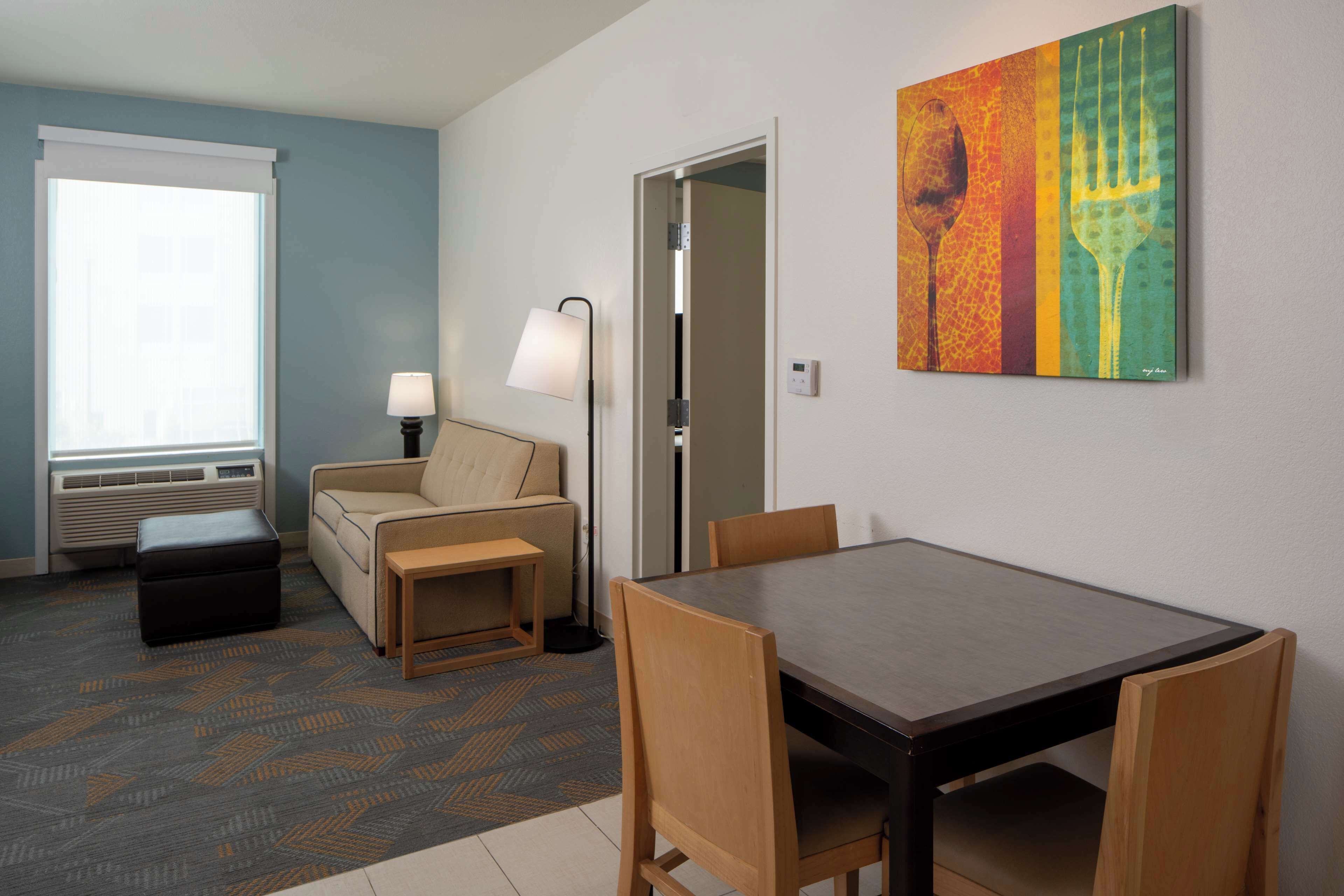 A hotel room with a dining table, chairs, a sofa, a lamp, and a painting on the wall.