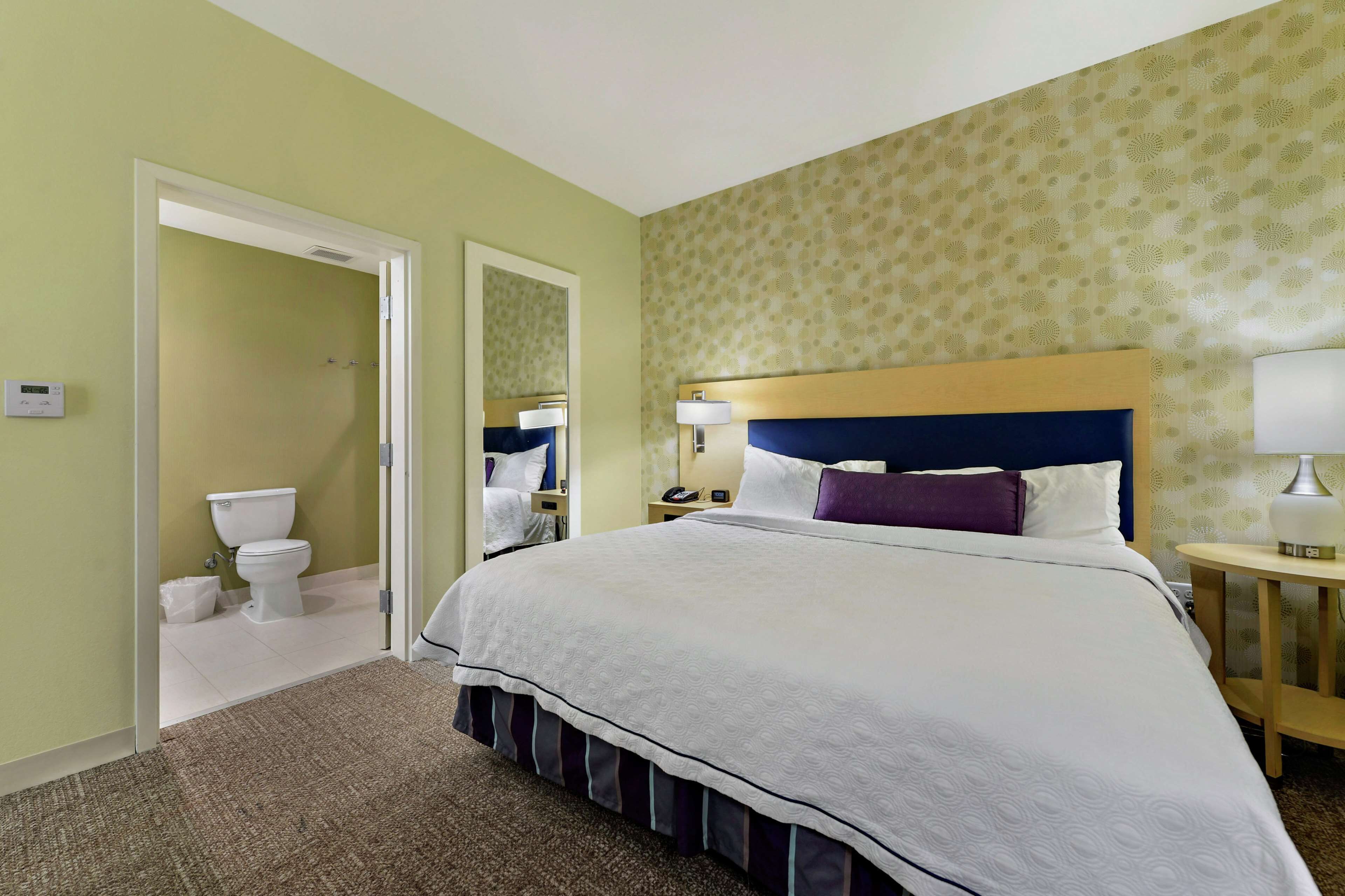 Una habitación de hotel con una cama grande, una mesita de noche con lámpara, un baño con inodoro y ducha, y un papel tapiz estampado.
