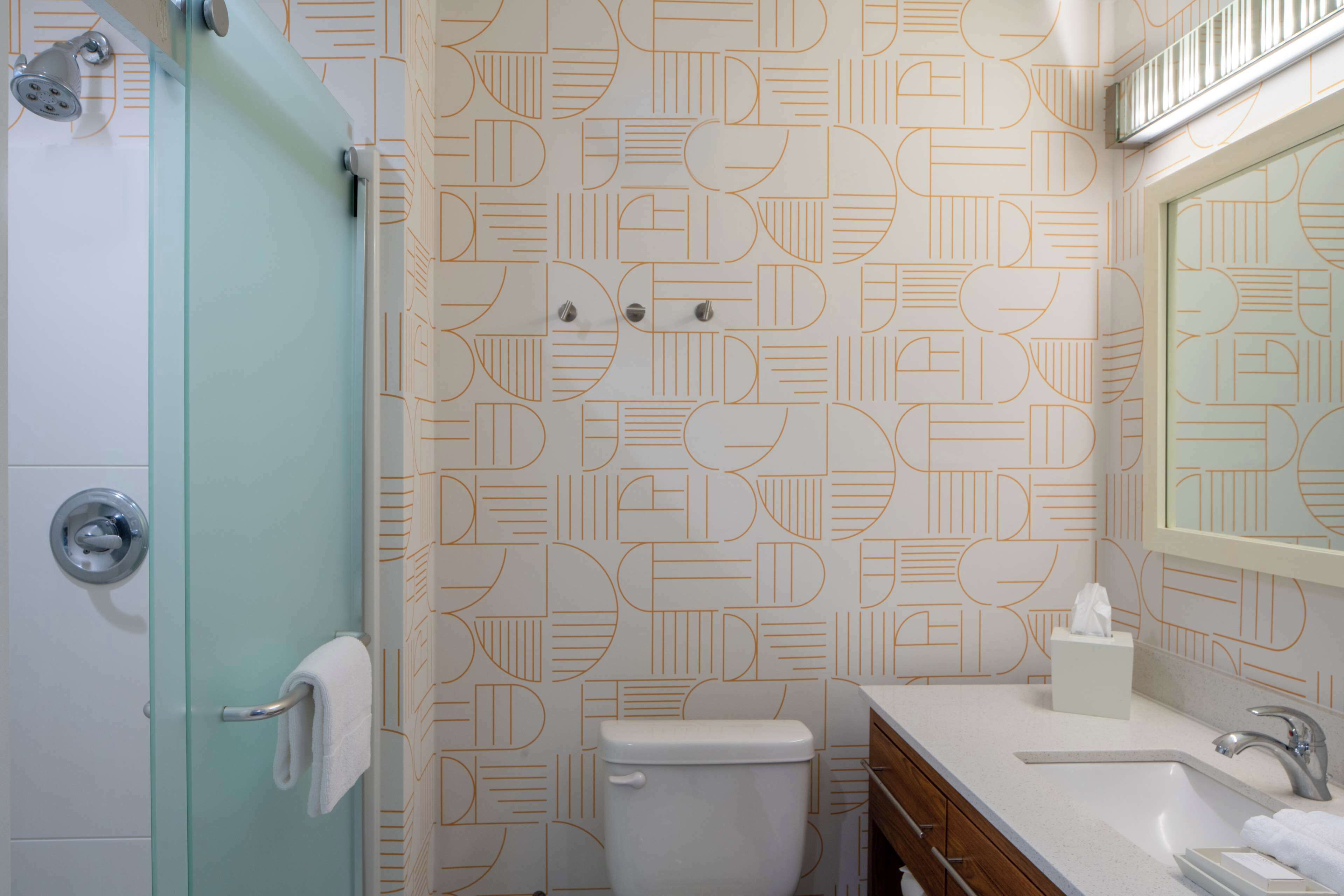 A bathroom with a geometric patterned wall, a glass shower enclosure, a white toilet, a sink with a mirror, and a towel rack.