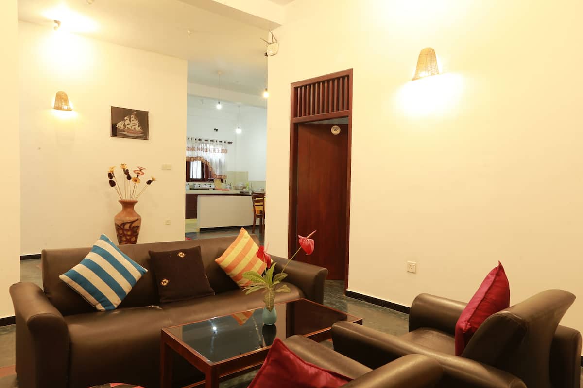 A living room with a sofa, coffee table, and a vase with flowers. There is a wooden door and a kitchen in the background.