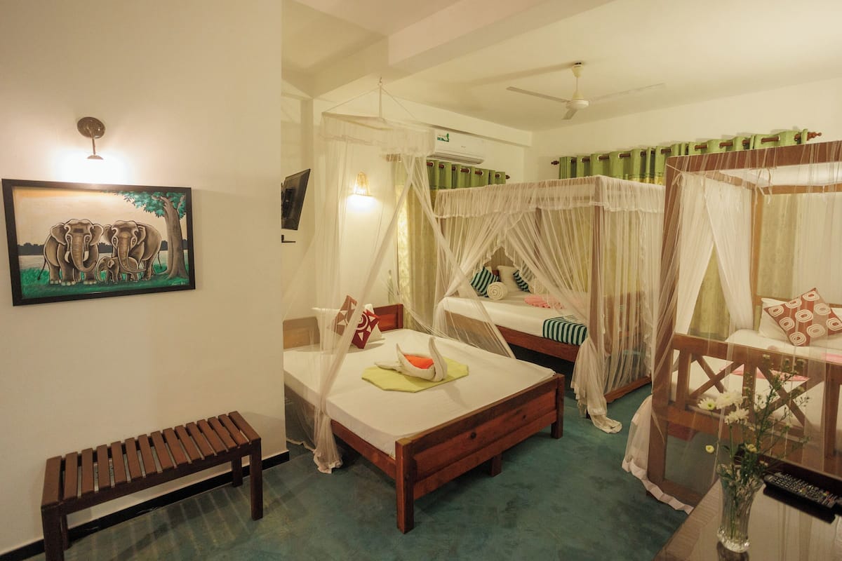 A room with two single beds, a bench, a bedside table, a wall-mounted TV, and a painting of elephants.