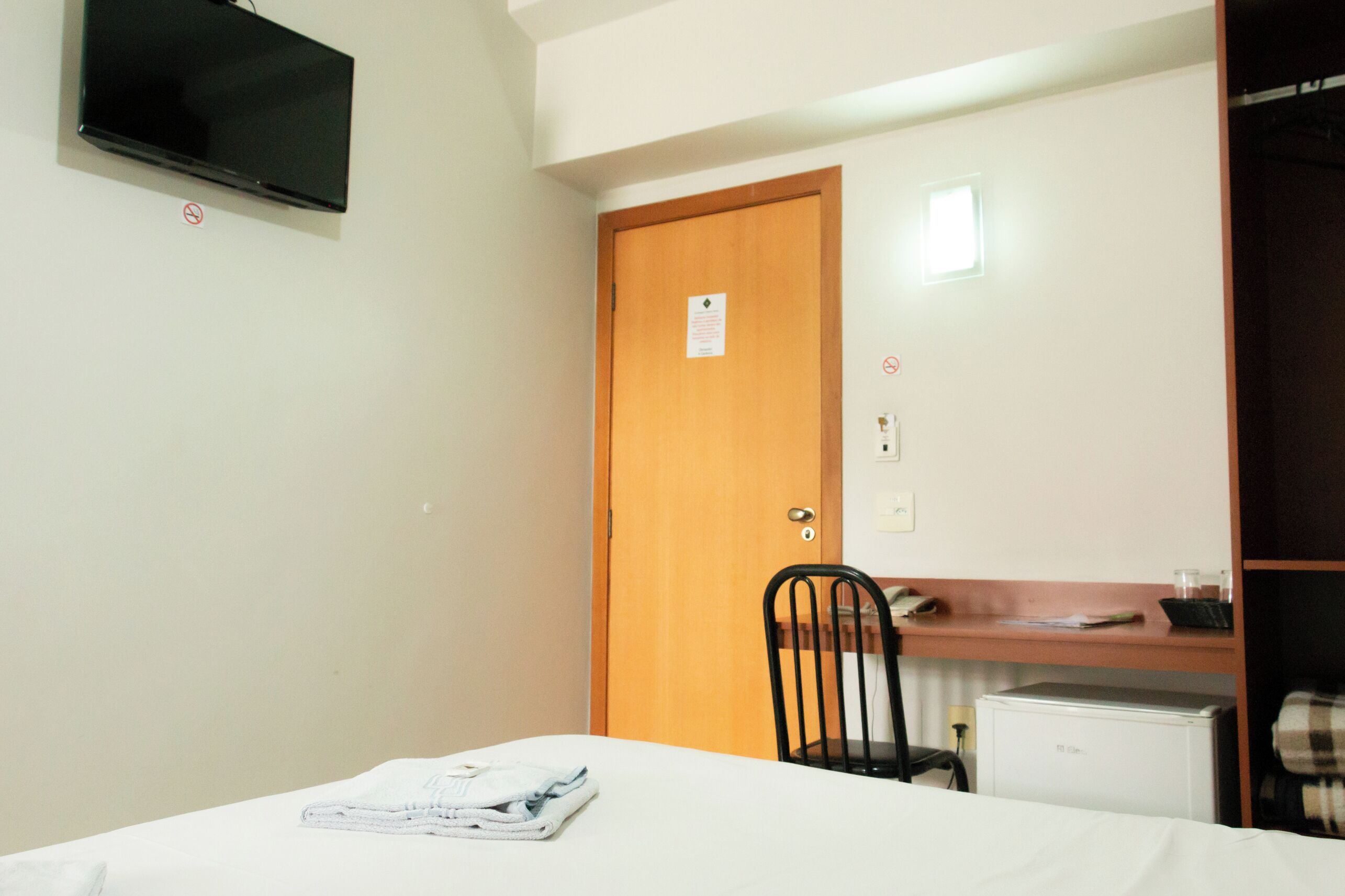 Habitación de hotel con cama, escritorio, silla, televisor montado en la pared y puerta con un letrero.