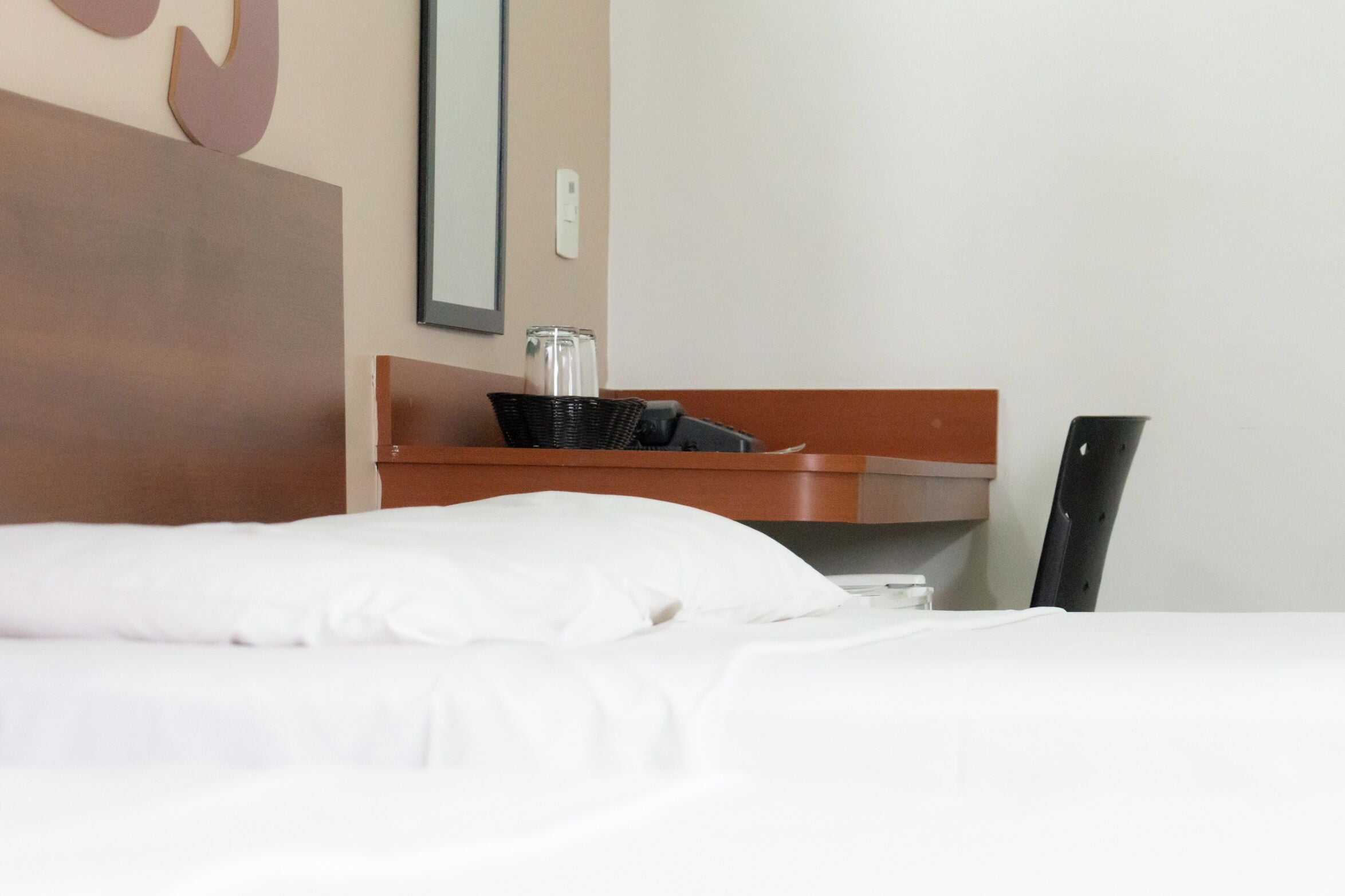Quarto de hotel com uma cama bem arrumada, cabeceira de madeira, pequena mesa lateral de madeira com um pote de vidro e uma cesta preta, e uma cadeira preta.