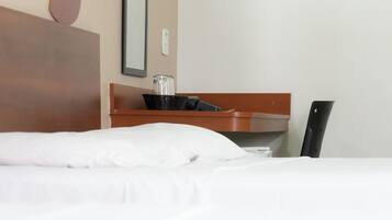 Quarto de hotel com uma cama bem arrumada, cabeceira de madeira, pequena mesa lateral de madeira com um pote de vidro e uma cesta preta, e uma cadeira preta.