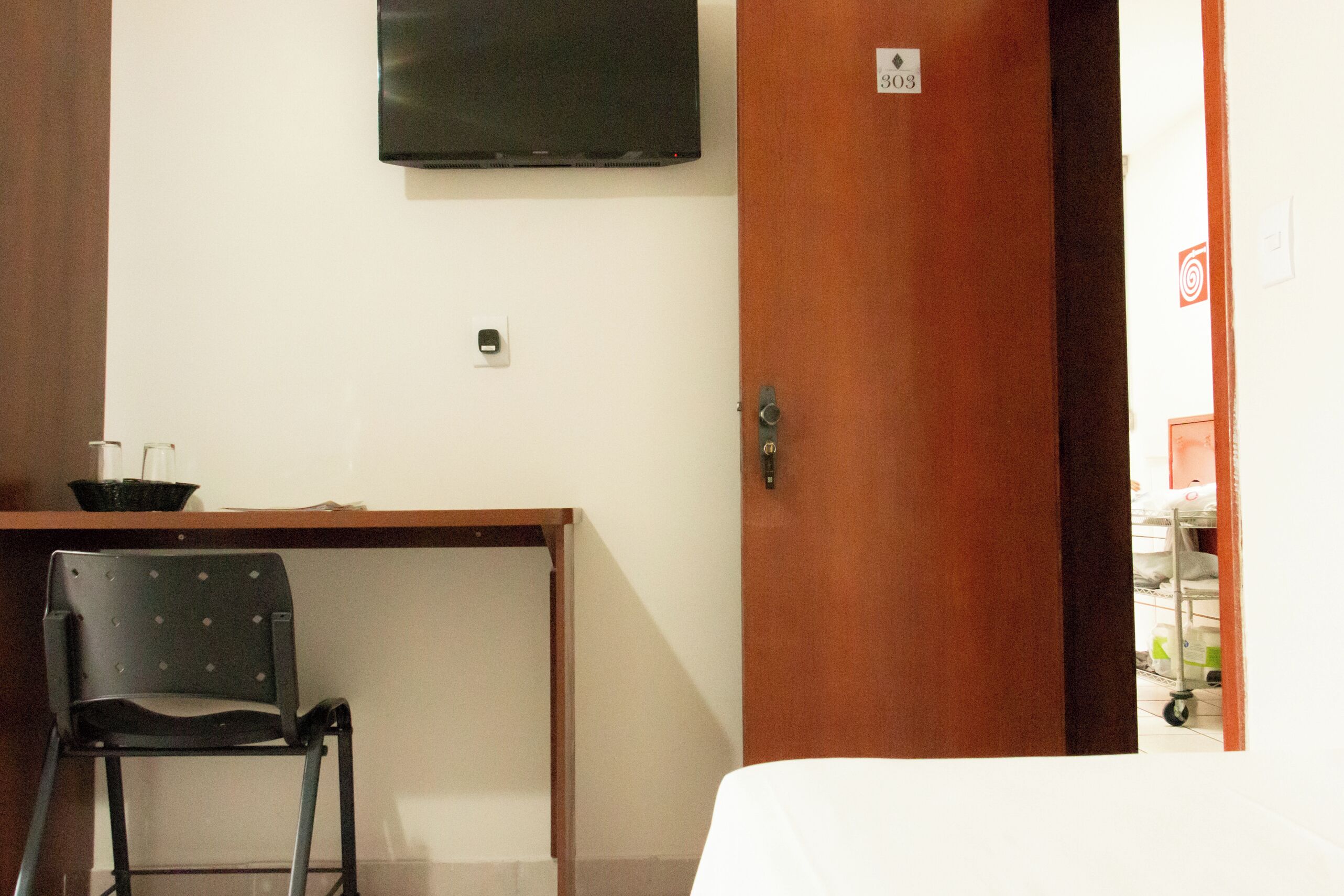 Una habitación con un escritorio de madera, una silla negra, una cama y un televisor montado en la pared.