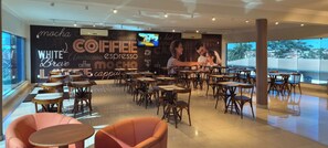 A coffee shop interior with tables and chairs, a large wall with coffee-related text, and a TV screen. - Bernal Hotel Econômico (Ituiutaba)
