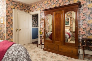 A bedroom with floral wallpaper, a wooden wardrobe, a bed with a floral bedspread, and a small table with a vase of flowers. - The Livingston Inn (Madison)