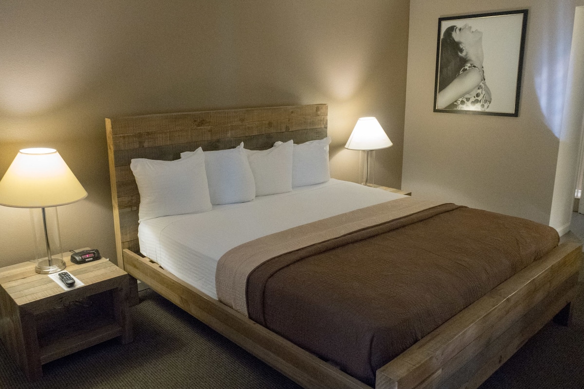 Cama bem arrumada com lençóis brancos e manta marrom, com duas mesas de cabeceira com luminárias, uma das quais tem um relógio digital.
