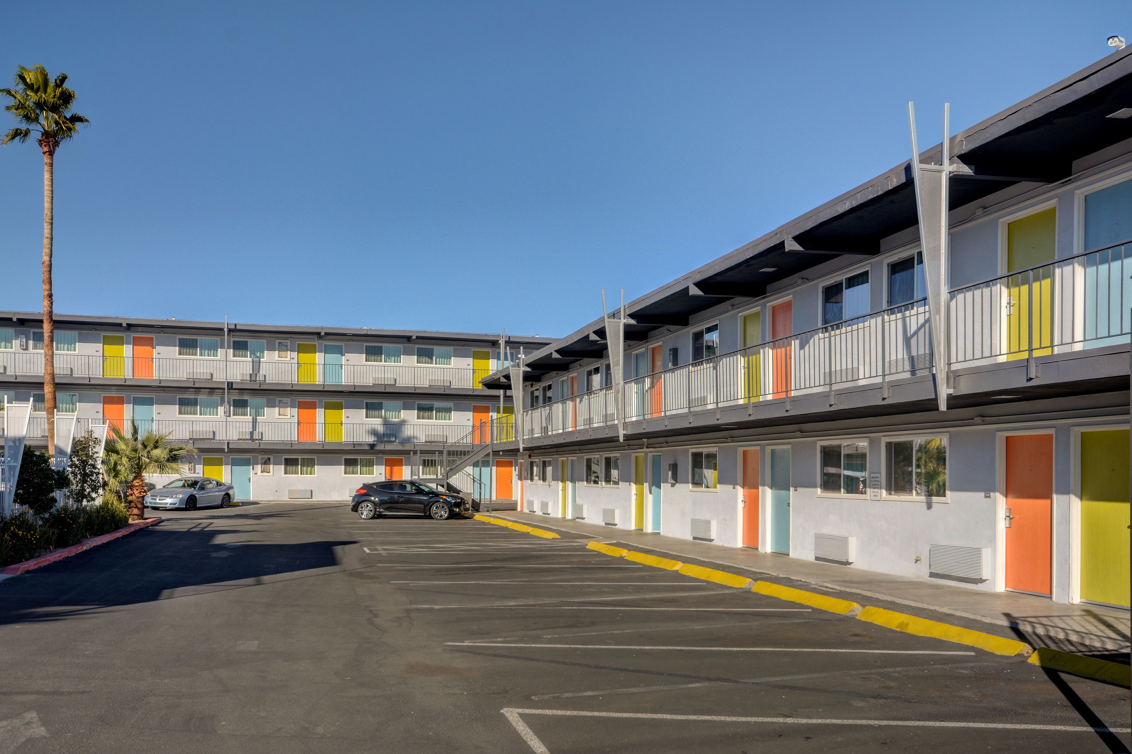 A multi-story motel with colorful doors and balconies, a parking lot with two cars, and a palm tree.