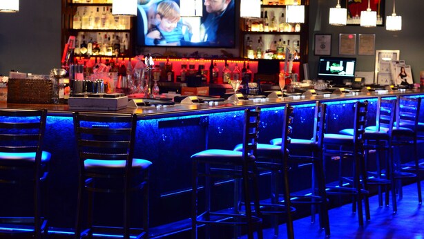 A bar area with multiple bar stools, a television screen, and a variety of bottles and glasses on shelves. - Thunderbird Hotel (Las Vegas)