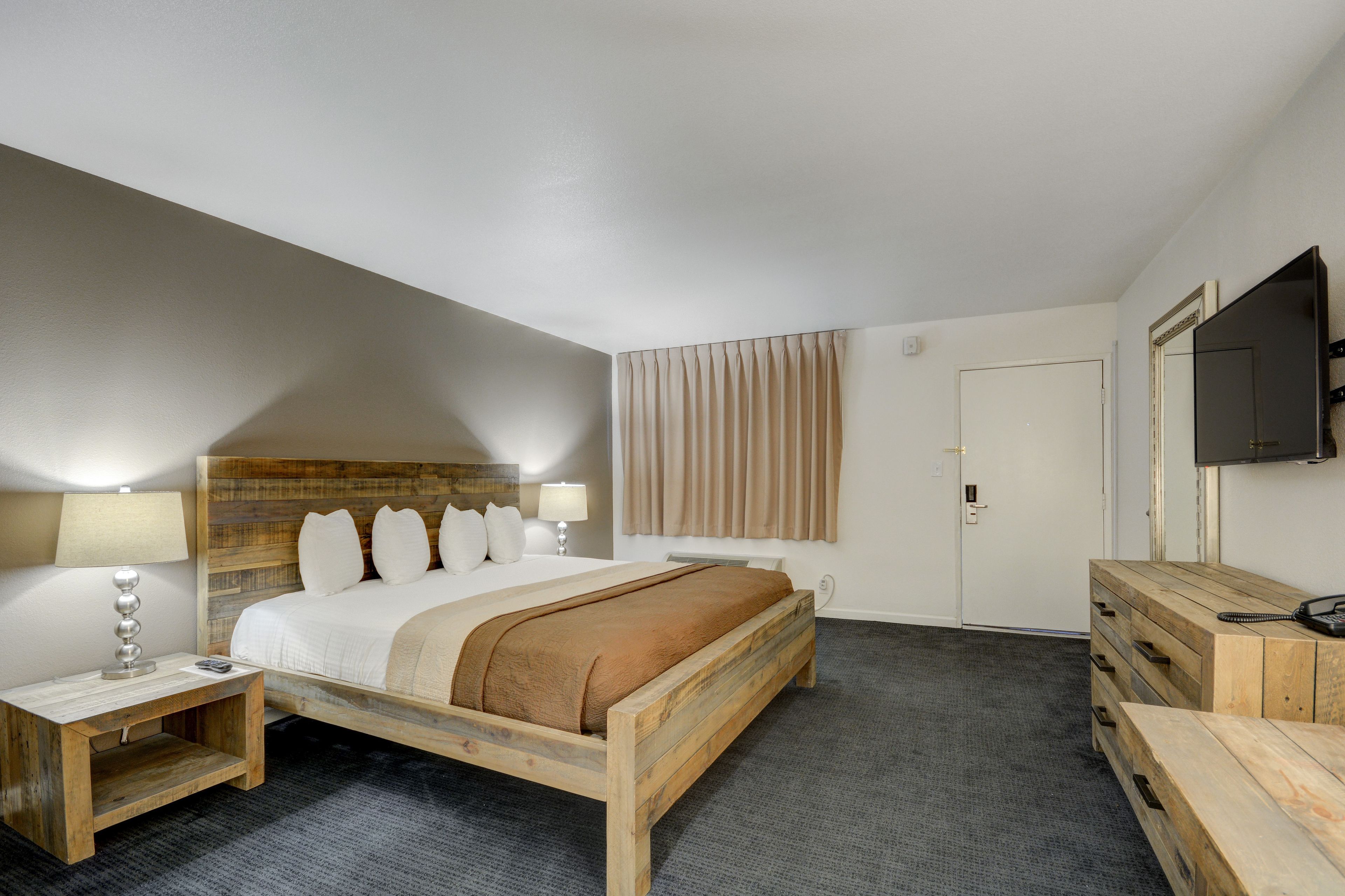 Camera d'albergo con un letto in legno, un comodino con una lampada, una cassettiera, una TV a parete e una porta con tenda.