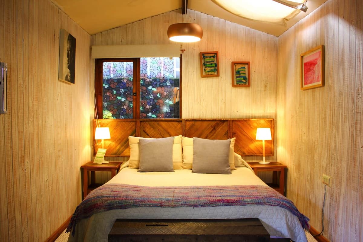 A bedroom with a wooden headboard, two bedside lamps, a bed with a colorful blanket, and a window with a stained glass design.